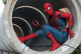 Spider-Man sitting inside a concrete pipe with his mobile phone and backpack.