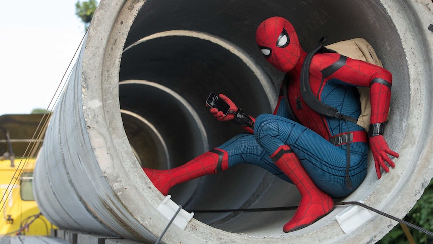 Spider-Man sitting inside a concrete pipe with his mobile phone and backpack.