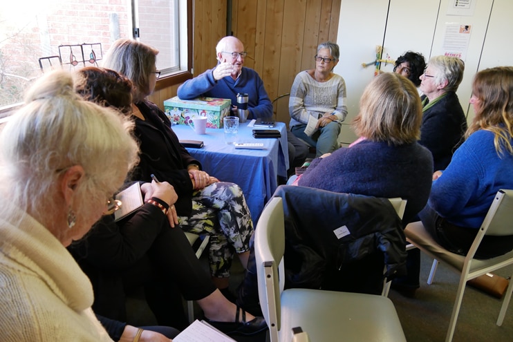 a room of women and one man sit in a circle talking to each other and writing on notepads