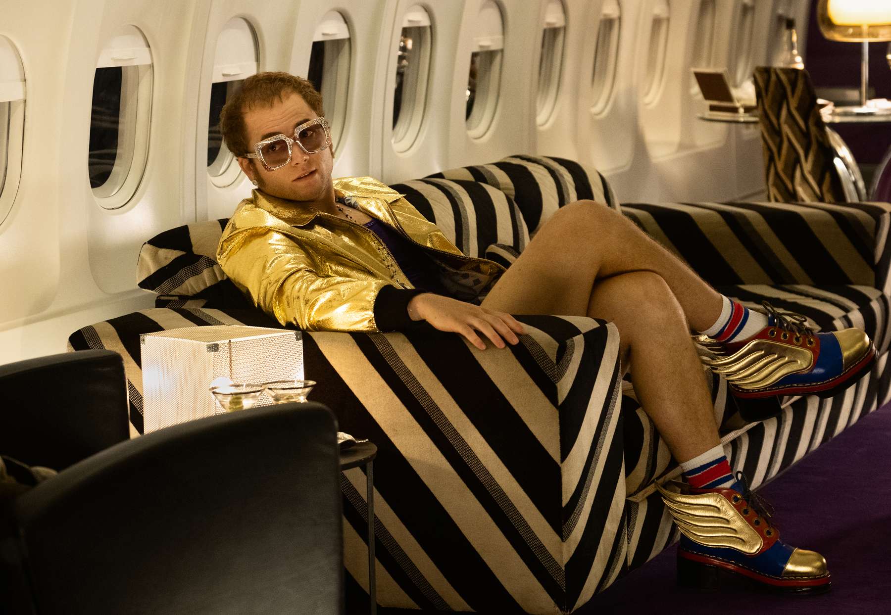Balding middle-aged man in gold leather jacket and bare legs wears big glasses, sits on coach in plane.