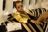 Balding middle-aged man in gold leather jacket and bare legs wears big glasses, sits on coach in plane.