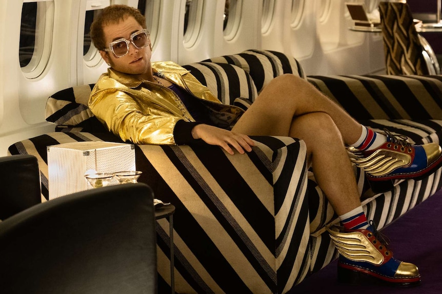 Taron Egerton, acting as Elton John, wears a gold leather jacket and big glasses while sitting on couch in a plane.