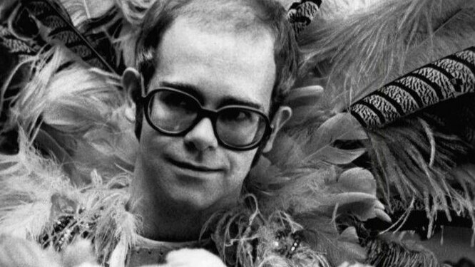 A black and white picture of Elton John wearing glasses and a feathery costume at the 1975 Rock Music Awards.