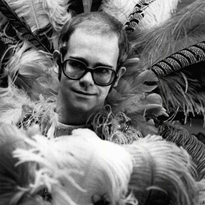 A black and white picture of Elton John wearing glasses and a feathery costume at the 1975 Rock Music Awards.