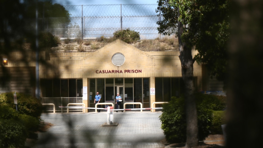 A wide shot of the front entrance to Casuarina Prison, with two men dressed in blue walking out and trees in the foreground.