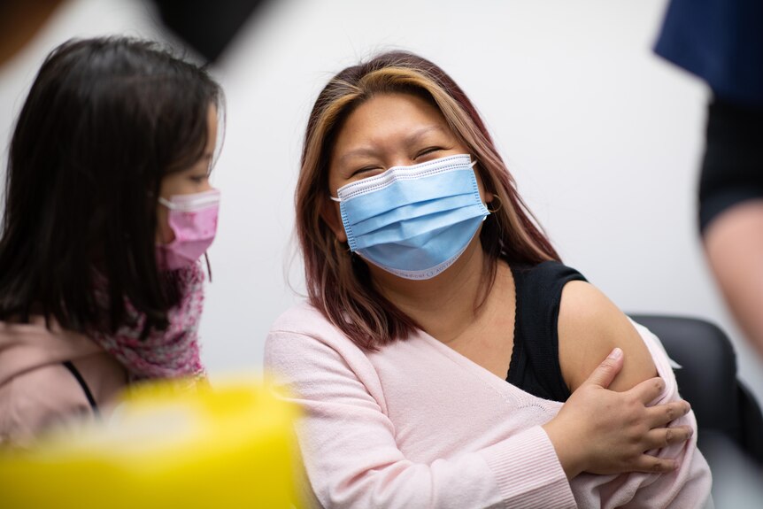A woman wearing a surgical mask holds her bare arm with her sleeve down. A young girl wearing a mask stands next to her