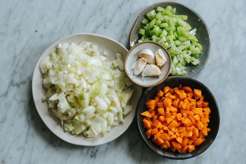 Bowls containing diced onion, diced carrot, chopped celery and garlic cloves