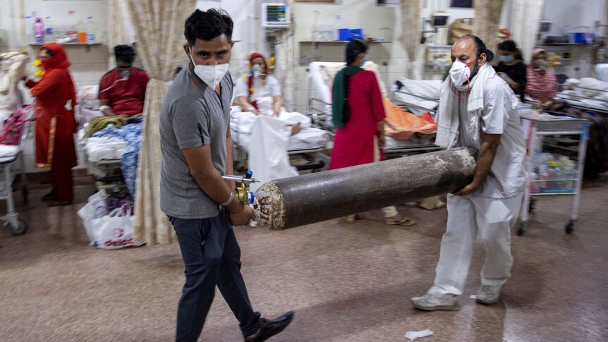 Two men wearing face masks carry an oxygen tank through a busy hospital ward.