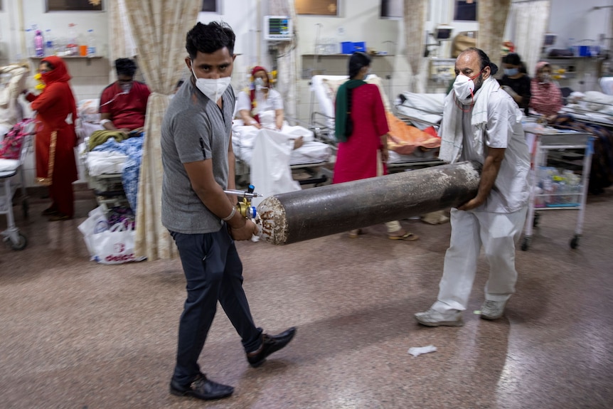 Two men wearing face masks carry an oxygen tank through a busy hospital ward.