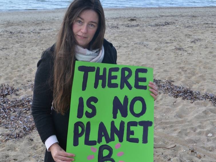Woman with long dark hair standing on the beach holding up a sign that says "there is no planet B"