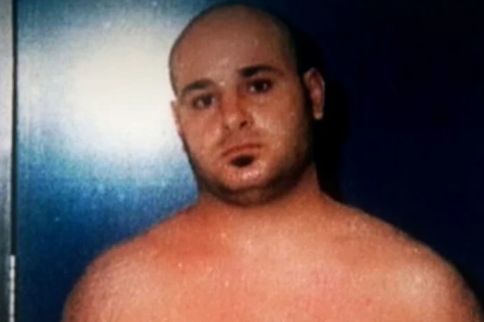 A man with a shaved head looks forward standing in front of a blue door