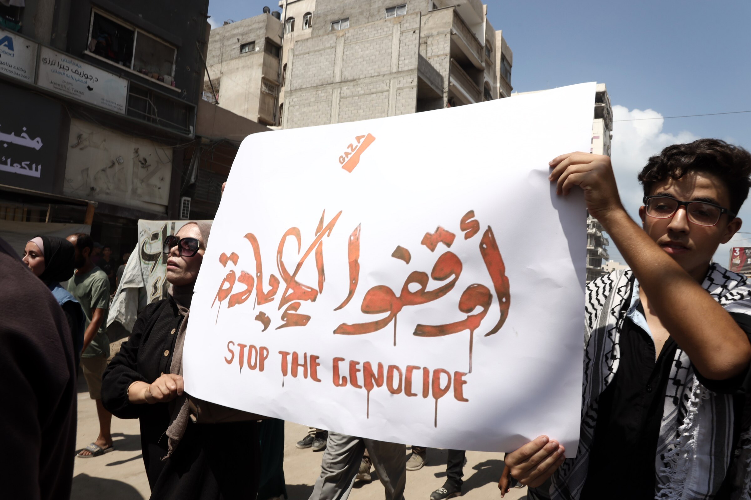 Gaza City protesters in the street holding a 'Stop the Genocide' sign.