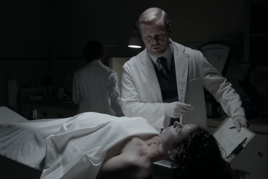 Craig McLachlan in a doctor's coat examines woman's dead body with sheet laying over her.