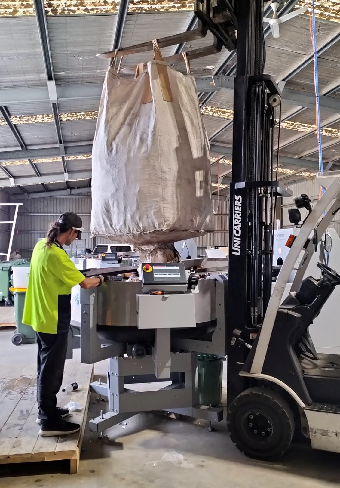 A man in high vis operates a forklift that is holding a big bag over the sorting machine.