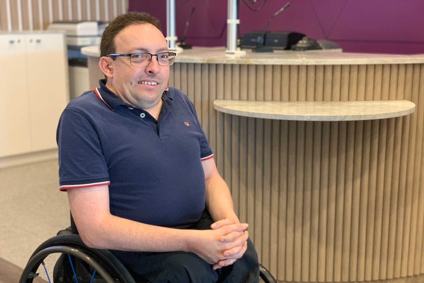 A man in a wheelchair with his hands clasped together in front of a desk at a bank Ausnew Home Care, NDIS registered provider, My Aged Care