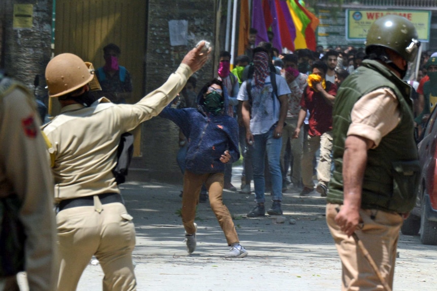 A young person wearing a hood and mask throws a rock towards security forces, who are wearing helmets.