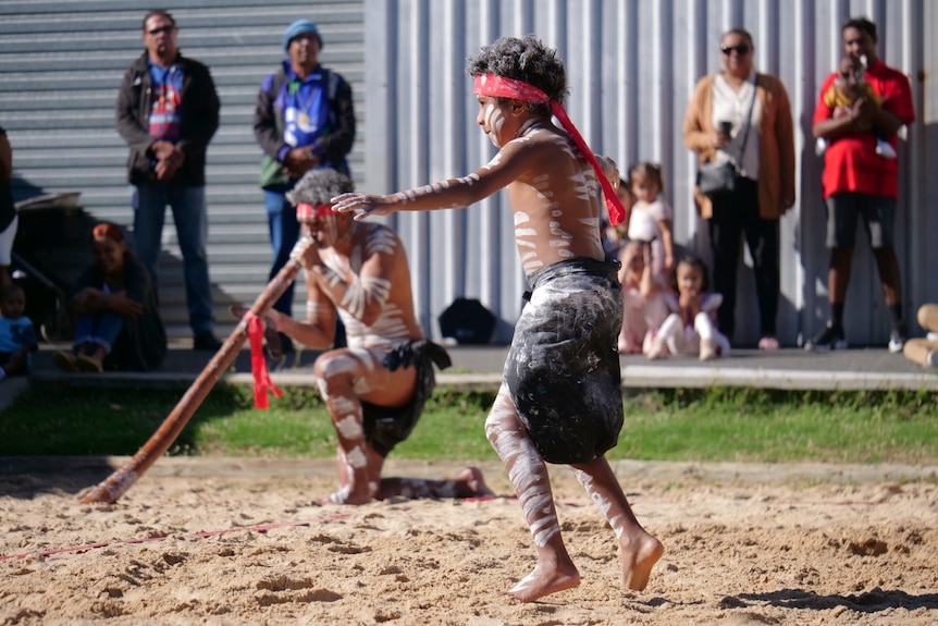 Walali dancing in traditional paint and black and red clothing, man playing didgeridoo in background, onlookers behind.