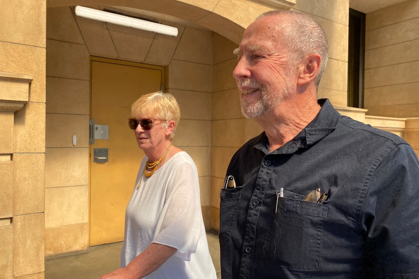 A woman with blonde short hair and a man with grey hair and a grey beard walk past an Adelaide court building.