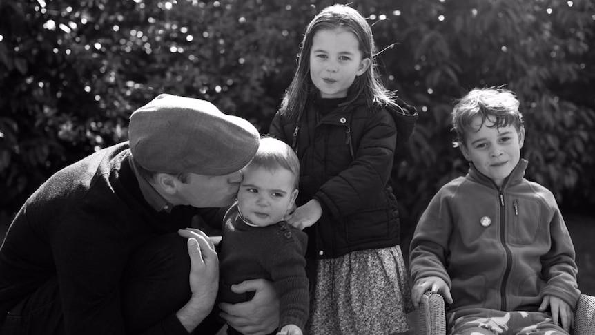 A black and white photo shows Prince William kissing a young Prince Louis while the other two children look at the camera.