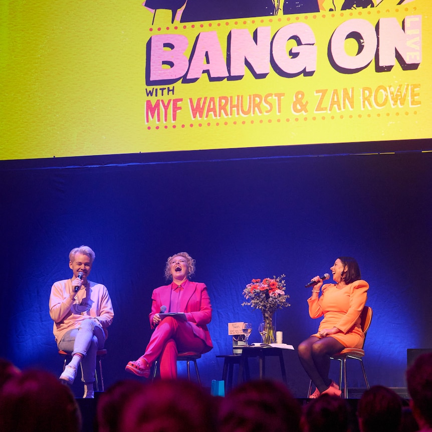 Joel, Zan and Myf sitting on stage under a Bang On Live poster with audience members watching in the foreground