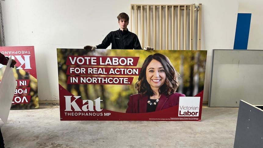 A man stands with a red-font and coloured billboard for Kat Theophanous.