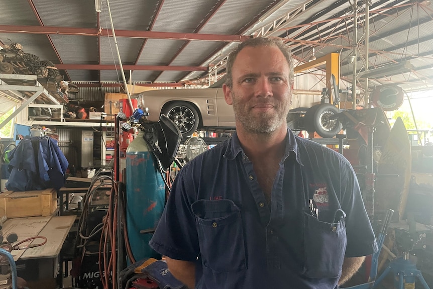 a mechanic stands in front of a raised vintage car in a workshop