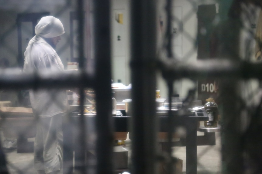 A detainee in white clothes and a white head scarf prepares a meal, viewed through glass or a fence which blurs the image
