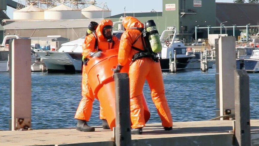 Firefighters in protective suits and wearing oxygen marks handle a large container on a jetty.