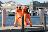 Firefighters in protective suits and wearing oxygen marks handle a large container on a jetty.