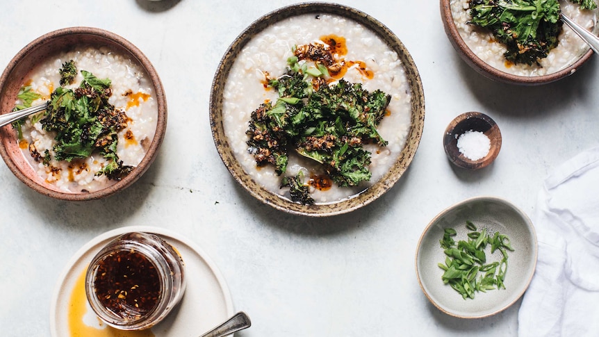 Three bowls of congee surrounded by assorted toppings including chilli oil, shallots, dried onion to accompany a recipe.