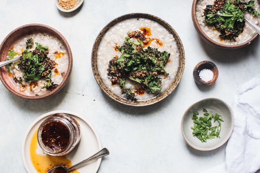 Three bowls of congee surrounded by assorted toppings including chilli oil, shallots, dried onion to accompany a recipe.