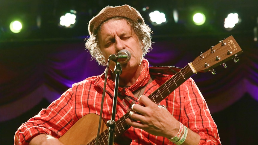 man wearing brown cap and red checkered shirt places acoustic guitar and sings on stage