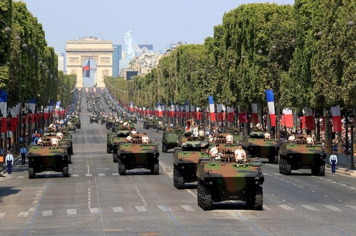 Countless tanks drive down the road with crowds gathered behind trees adorned with French flags in front of them.
