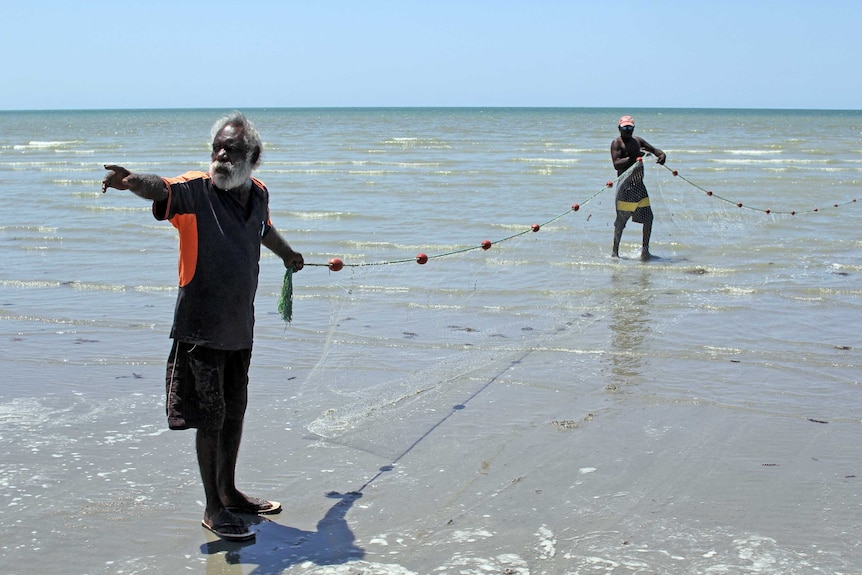 a man pointing and holding a net in the shallows of a beach with another man holding the net behind.