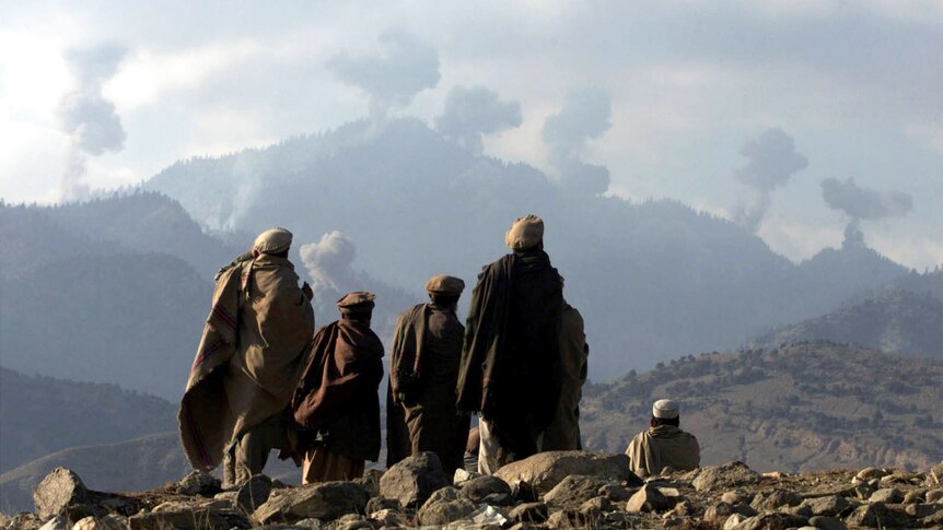 A group of people dressed in black thawb look at mountains with smoke rising in the distance.