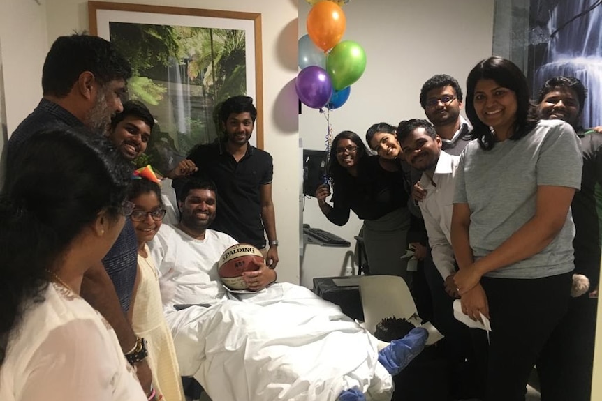 Ishan Jayasekara smiles in his hospital bed, surrounded by smiling people, holding a basketball. There are balloons behind him.