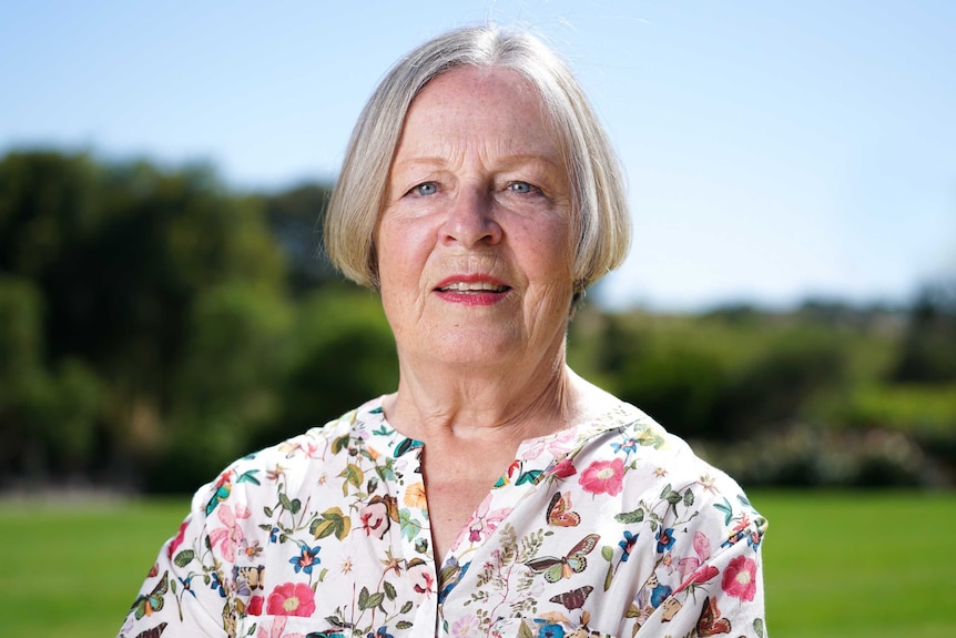 A woman with short grey hair looks directly at the camera in front of an outdoor backdrop of greenery and blue sky.