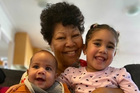 Aunty Violet Langan smiles as she holds a baby on her left and a toddler on her right while sitting on a sofa.