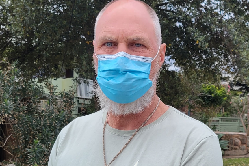 An older man, balded-headed and bearded, stands wearing a mask out the front of his coastal home.