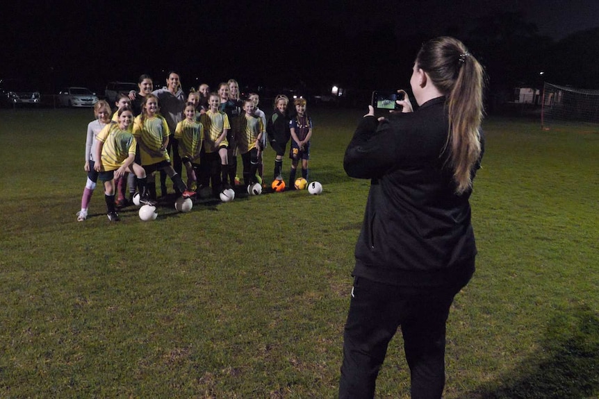 A dozen girls in football uniforms pose for photos at a soccer ground