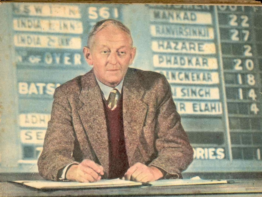 A man sits at a desk in front of an old cricket scoreboard