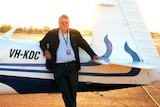 An man wearing glasses is standing in front of a plane with the sun setting behind him