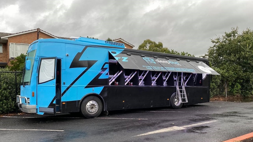 A blue and black bus with the side lifted e to reveal a ladder and sleep pods inside