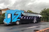 A blue and black bus with the side lifted e to reveal a ladder and sleep pods inside