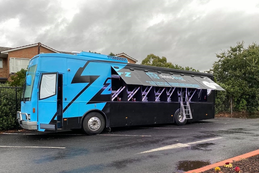 A blue and black bus with the side lifted e to reveal a ladder and sleep pods inside