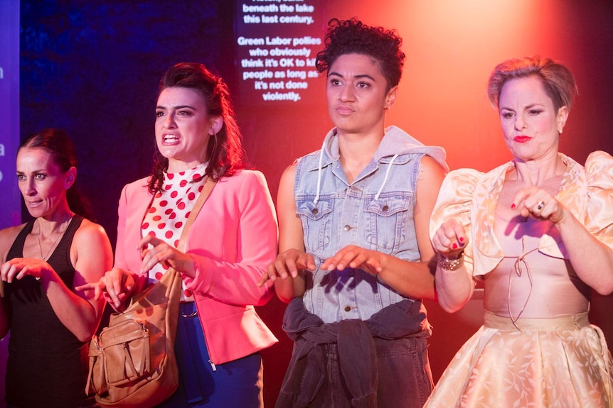 Four women stand on stage with hands in front of them as if typing, with text about Australian politics on a screen behind them.