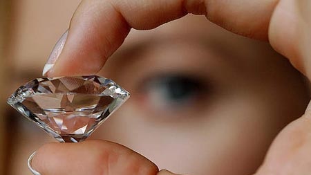 Close-up of a large diamond held between two fingers, with background of a blurred eye