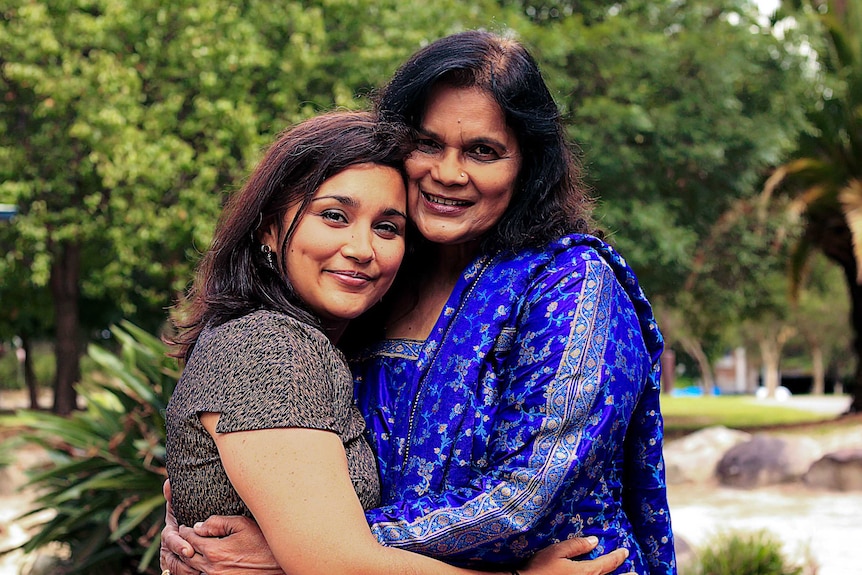 Two women embracing for a photo standing in front of green trees