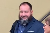 A man with a voluminous beard and dark, close-cropped hair, wearing a blazer and smiling.