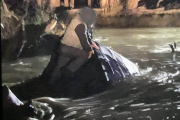 A man clings to part of a vehicle, with the rest submerged in floodwater.